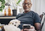 Smiling retired senior male using smart phone while sitting with dog in room at home