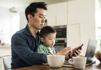 Father multi-tasking with young son (2 yrs) at kitchen table