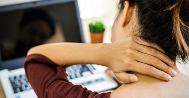 Woman sitting in front of computer rubbing the back of her neck in pain.
