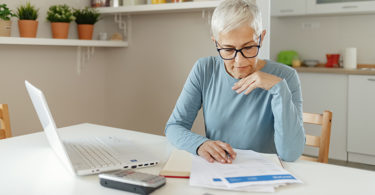 Senior woman working on home finance. Mature woman checking bills at home. Older woman checking pension approval certificate with laptop at home.
