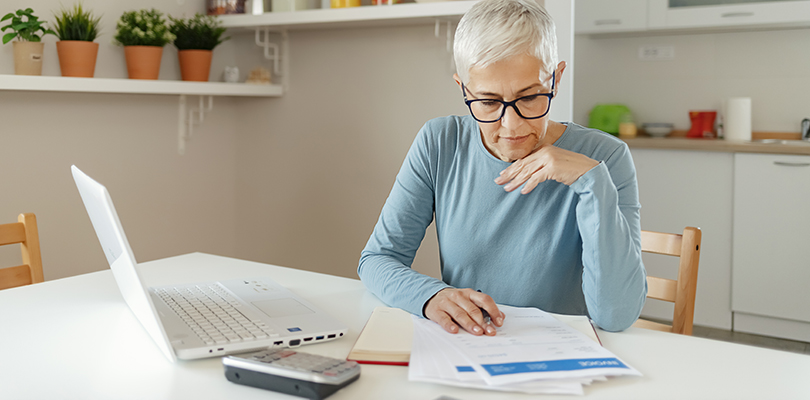 Senior woman working on home finance. Mature woman checking bills at home. Older woman checking pension approval certificate with laptop at home.