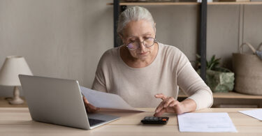 Concentrated old woman in eyeglasses calculating utility bills or domestic expenditures, doing financial paperwork, paying for services or insurance using computer e-banking application alone at home.