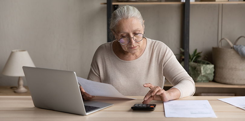 Concentrated old woman in eyeglasses calculating utility bills or domestic expenditures, doing financial paperwork, paying for services or insurance using computer e-banking application alone at home.