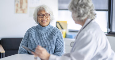 A senior patient of Asian decent, sits across the desk from her doctor as they review her recent tests results. The doctor is wearing a white lab coat and holding a tablet out in her hands as the patient looks at the screen and listens attentively.