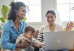 Young mothers with baby girl in living room. One woman using laptop for work or online shopping.