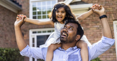 Toddler daughter riding on father's shoulders in front of home