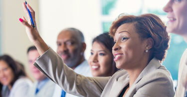 Mid adult African American businesswoman is smiling and raising her hand. She is answering a question during a business conference or job training seminar. Professional woman is wearing business clothing. She is sitting with diverse colleagues.