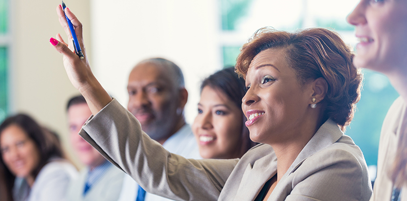 Mid adult African American businesswoman is smiling and raising her hand. She is answering a question during a business conference or job training seminar. Professional woman is wearing business clothing. She is sitting with diverse colleagues.