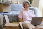 Middle-aged woman sitting in room with using laptop surrounded by moving boxes.