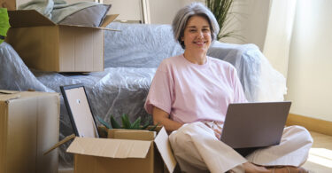 Middle-aged woman sitting in room with using laptop surrounded by moving boxes.