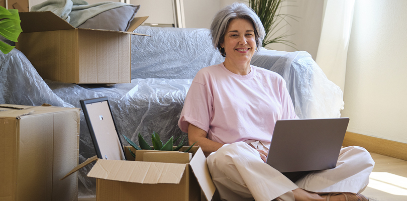 Middle-aged woman sitting in room with using laptop surrounded by moving boxes.