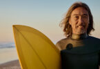 Smiling senior holds surfboard and stands next to rising sun