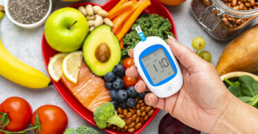 Hand holding glucose monitor above plate of food including salmon, broccoli, blueberries, and avocado