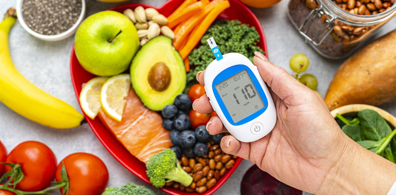 Hand holding glucose monitor above plate of food including salmon, broccoli, blueberries, and avocado