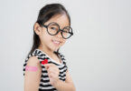 Smiling young girl wearing glasses with a bandaid on her upper arm holding a red heart sticker.