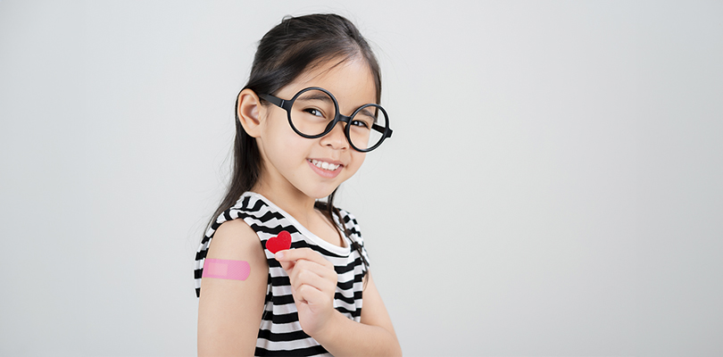 Smiling young girl wearing glasses with a bandaid on her upper arm holding a red heart sticker.