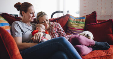 Same sex couple enjoying a cuddle together at home, with their baby daughter and their pet dog.