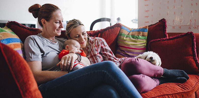 Same sex couple enjoying a cuddle together at home, with their baby daughter and their pet dog.