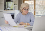 Woman wearing glasses holding document looking at laptop.