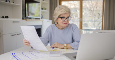 Woman wearing glasses holding document looking at laptop.