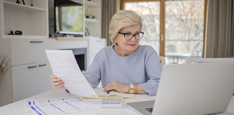 Woman wearing glasses holding document looking at laptop.