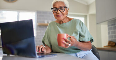 Senior Black woman holding mug in one hand using laptop with the other