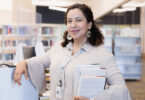 The mid adult female head librarian of the public library smiles as she stands by the checkout desk.