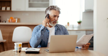Older man on phone sitting in front of table with laptop