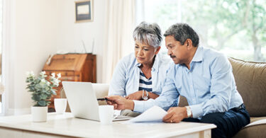Two seniors sitting on couch looking at laptop, one is holding papers and pointing at laptop screen with a pen