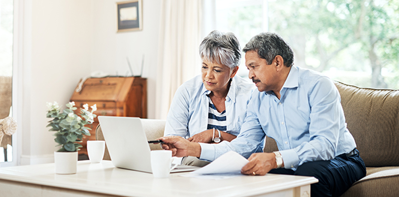Two seniors sitting on couch looking at laptop, one is holding papers and pointing at laptop screen with a pen